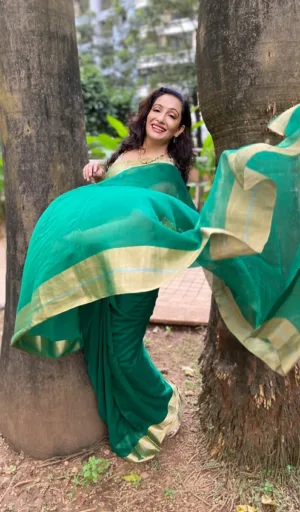 Woman wearing a green saree with a gold border, playfully posing outdoors near trees.