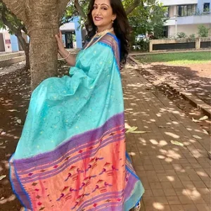 Woman wearing a turquoise handloom Paithani saree with Bandhani-style motifs and peach–purple border, posing outdoors near a tree; elegant traditional festive saree look.